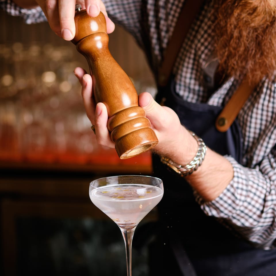 Bartender grinds a wooden pepper mill over a clear cocktail garnished with floating citrus peels at a bar.