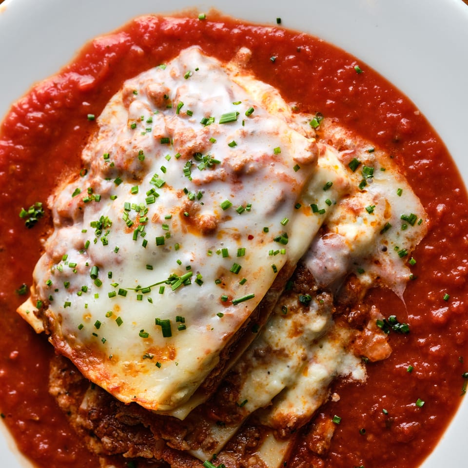 Layered lasagna topped with melted cheese and chives, surrounded by tomato sauce on a white plate.