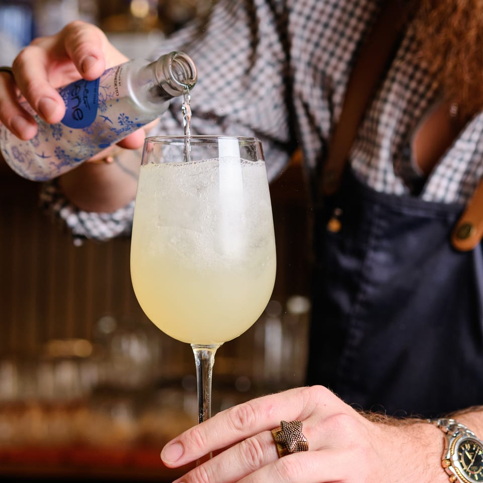 Bartender pours a clear spirit from a decorated bottle into a frothy pale cocktail at a warmly lit bar.