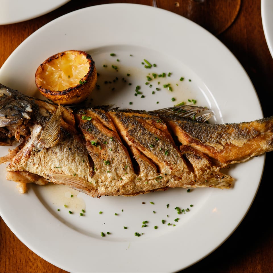 Whole grilled fish served with a charred lemon half and chopped herbs on a white plate.