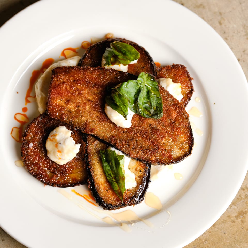 Crispy fried eggplant slices layered with creamy sauce, fresh basil, and drizzled sauces on a white plate.