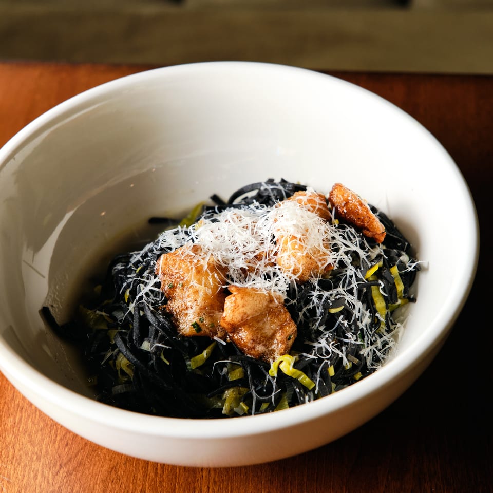 White bowl of black pasta topped with grilled salmon, shredded garnish, and green vegetables on a wooden restaurant table.