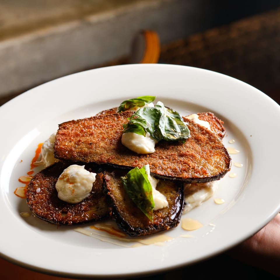 Crispy breaded eggplant slices layered with creamy sauce, fresh greens, and drizzled oils on a white plate.