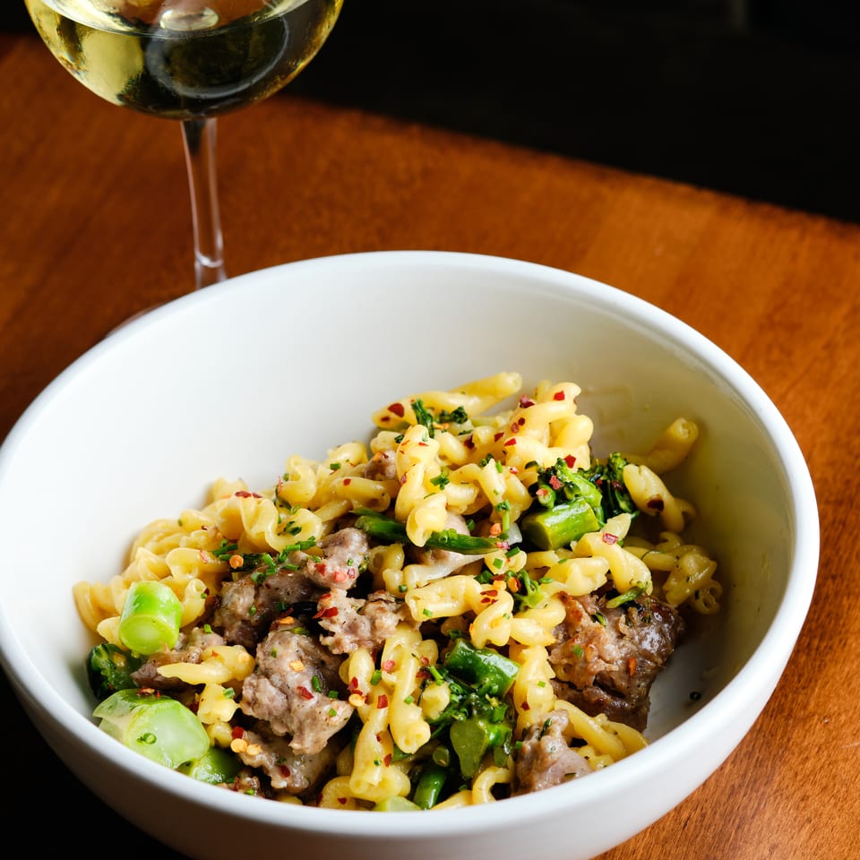 Bowl of pasta with sausage, green vegetables, herbs, and chili flakes beside a glass of red wine on a wooden table.