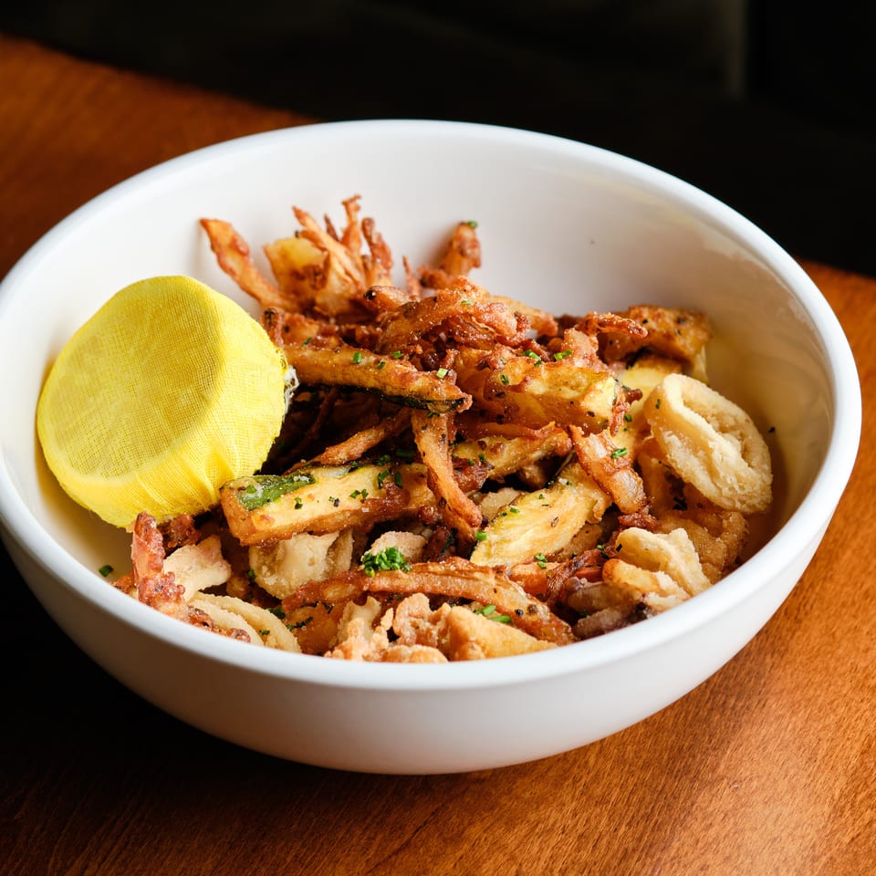 Crispy fried vegetable pieces garnished with herbs and lemon in a white bowl on a wooden table.
