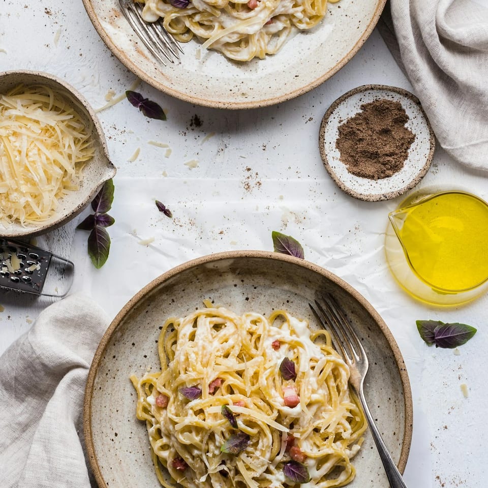 Two plates of creamy carbonara pasta with bacon, basil, and grated cheese on a white surface.